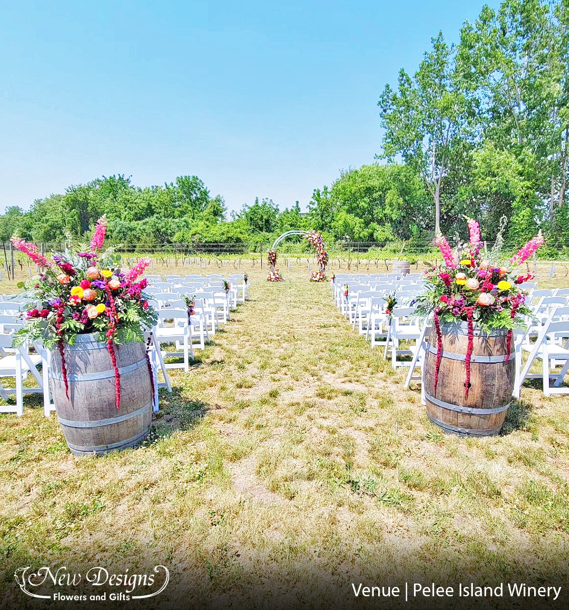 A Wedding Aisle in a Vinyard, At the head sits a White Metal Arch with 4 offset floral installments. Floral installments line the center chairs, and closest to the viewer sits 2 more large floral installments atop Wine Barrels. The flowers are wild and overflowing in various summer shades of Orange, Yellow. Pink. Purple and Blue